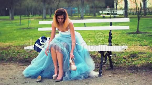 穿着银色和蓝色连衣裙的漂亮女孩坐在公园路的长凳上穿上高跟鞋看着谈话视频的预览图