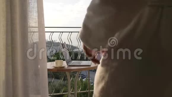 一个穿着白色实验室外套的人在俯瞰海湾的露台上的笔记本电脑上工作4K视频的预览图
