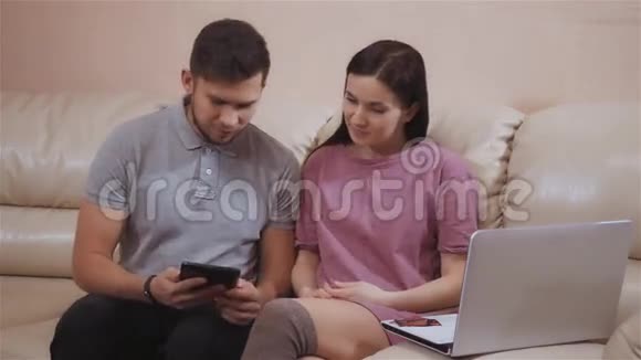 可爱的年轻夫妇使用笔记本电脑和平板电脑在家坐在皮革沙发上网上购物与塑料卡片视频的预览图
