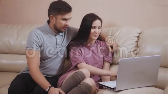 可爱的年轻夫妇使用笔记本电脑在家坐在皮革沙发上使用Skype视频的预览图