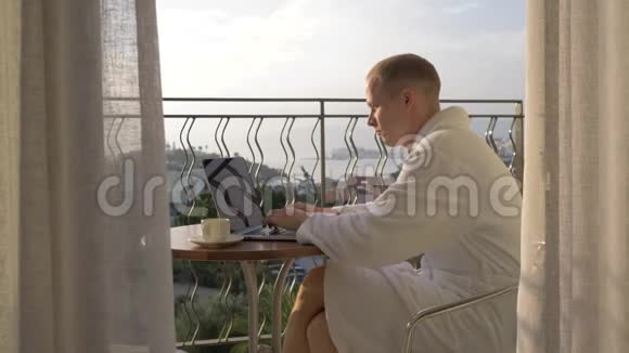 一个穿着白色实验室外套的人在俯瞰海湾的露台上的笔记本电脑上工作4K视频的预览图