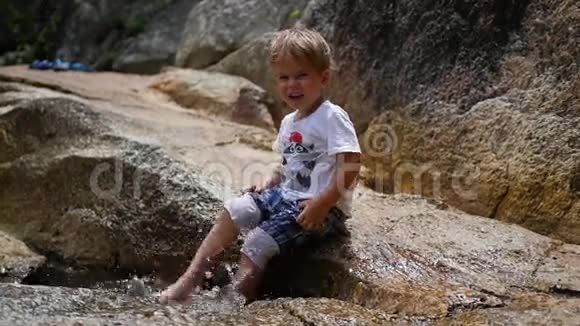 快乐的孩子坐在岩石上用脚溅起水花视频的预览图