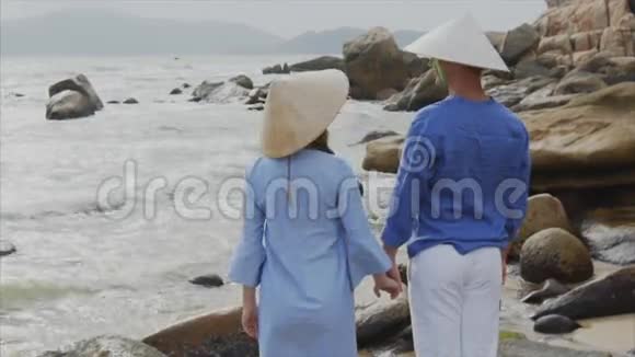 一对年轻漂亮的情侣戴着越南帽子穿着蓝色衣服手牵着手望着海滩视频的预览图