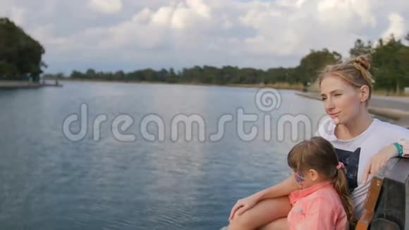年轻的母亲和小女孩在湖边的公园里享受阳光的夜晚坐在长凳上视频的预览图