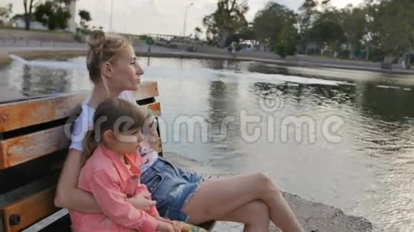 年轻的母亲和小女孩在湖边的公园里享受阳光的夜晚坐在长凳上视频的预览图