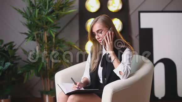 戴眼镜的年轻美女坐在办公室里打电话在笔记本上说和写上面的植物视频的预览图