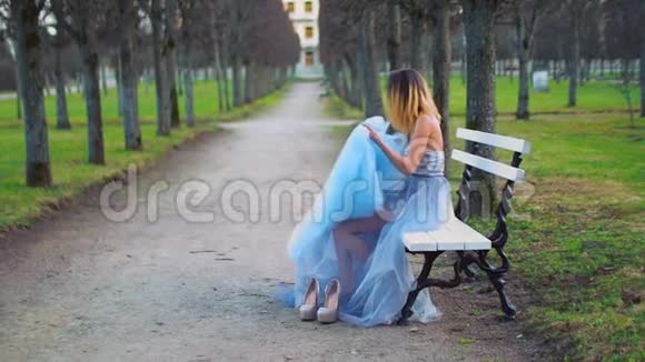 穿着银色和蓝色连衣裙的漂亮女孩坐在Parkway的长凳上调整着衣服穿上蓝色的长裙开始穿起来视频的预览图