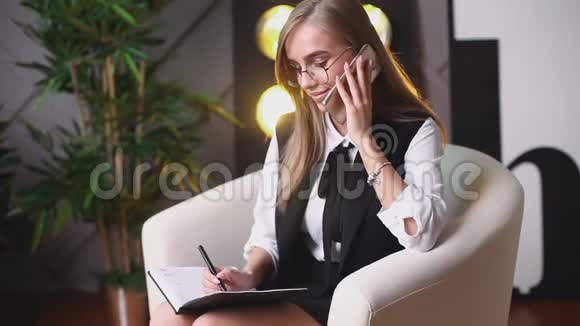 戴眼镜的年轻美女坐在办公室里打电话在笔记本上说和写上面的植物视频的预览图
