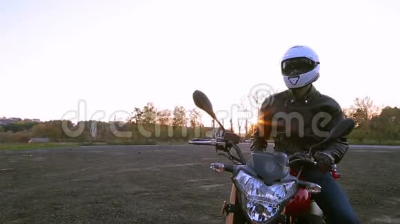 一个穿着黑色皮夹克和白色头盔的年轻人坐在摩托车上在秋日日落前开灯视频的预览图