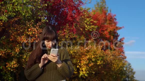 一位穿着绿色毛衣的年轻女子在秋天公园里使用智能手机视频的预览图