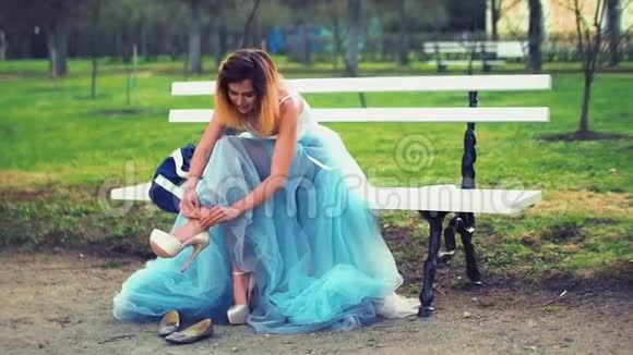 穿着银色和蓝色连衣裙的迷人女孩坐在帕克威的长凳上穿上高跟鞋摆出姿势站起来视频的预览图