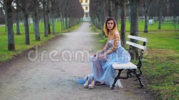 穿着银色和蓝色连衣裙的迷人女孩坐在公园路的长凳上摆姿势做脸在时玩得很开心视频的预览图