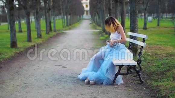 穿着银色和蓝色连衣裙的漂亮女孩坐在公园路的长凳上换衣服并准备在视频的预览图