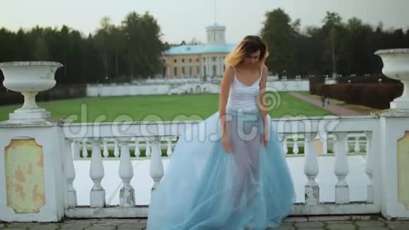 漂亮的瘦女孩穿着白色和蓝色的连衣裙站在白石栏杆附近在照片拍摄时微笑和摆姿势视频的预览图