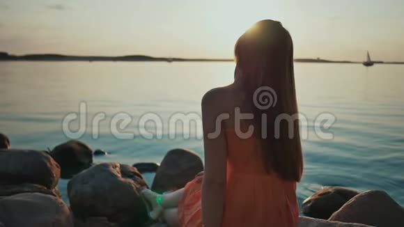 一个年轻的女孩坐在海滩上的岩石上周围没有人日落在潮湿的沙子和海水中反射关于视频的预览图