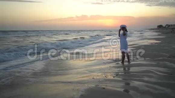 年轻漂亮的女人在日落时站在海边穿着白色连衣裙和白色帽子视频的预览图
