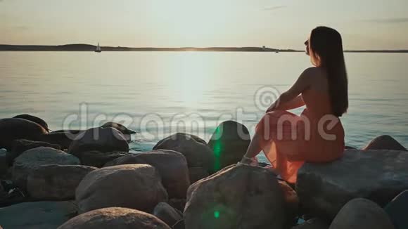 一个年轻的女孩坐在海滩上的岩石上周围没有人日落在潮湿的沙子和海水中反射关于视频的预览图