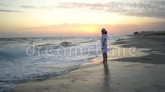 年轻漂亮的女人在日落时站在海边穿着白色连衣裙和白色帽子视频的预览图