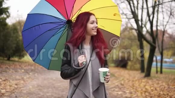 一位留着红发的年轻女子在秋天公园散步一边拿着五颜六色的雨伞一边从纸杯里喝着咖啡视频的预览图