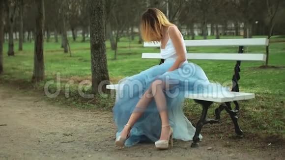 穿着银色和蓝色连衣裙的漂亮女孩坐在帕克威的长凳上穿着高跟鞋膝盖磕磕绊绊视频的预览图