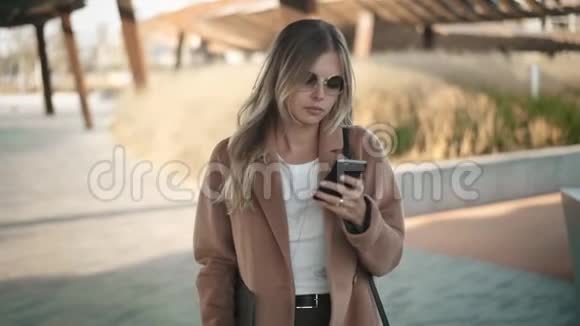 秋天穿着外套的漂亮金发女人走着看着她的智能手机视频的预览图