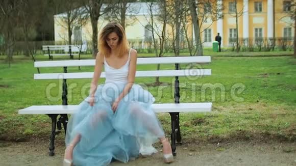漂亮的瘦女孩穿着银色和蓝色的连衣裙穿着高跟鞋坐在Parkway的长凳上膝盖敲着盖着盖子视频的预览图