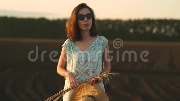 戴太阳镜的女孩站在日落时的田野里拿着金色的麦穗和草帽乡村户外场景视频的预览图