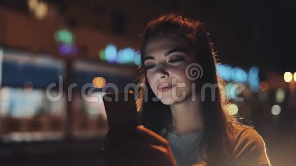 迷人的年轻女性使用智能手机站在夜城的街道上对这条信息做出了愉快的反应现代视频的预览图