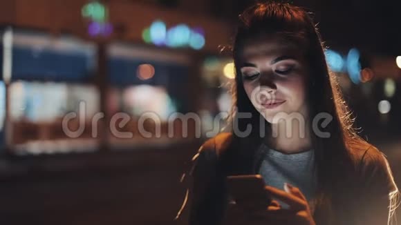 迷人的年轻女性使用智能手机站在夜城的街道上对这条信息做出了愉快的反应现代视频的预览图