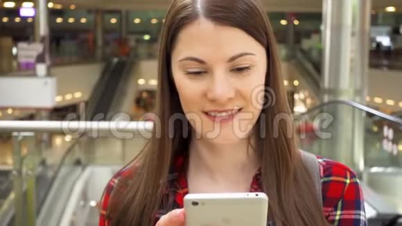 年轻漂亮的女人站在购物中心微笑用她的智能手机和朋友聊天视频的预览图