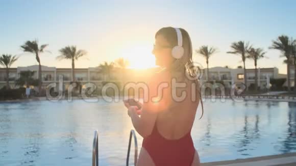 美丽的女人穿着红色泳衣和耳机在智能手机上跳舞和听音乐比基尼女郎的肖像视频的预览图