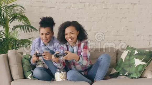 慢动作两个混合种族卷曲女孩朋友坐在沙发上玩控制台电脑游戏与游戏本和乐趣视频的预览图