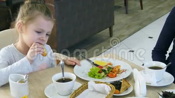 美丽的黑发母亲说服她年轻的金发女儿在咖啡馆吃蔬菜沙拉视频的预览图