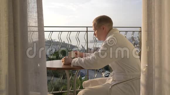一个穿着白色长袍的年轻人在露台上喝咖啡4K视频的预览图