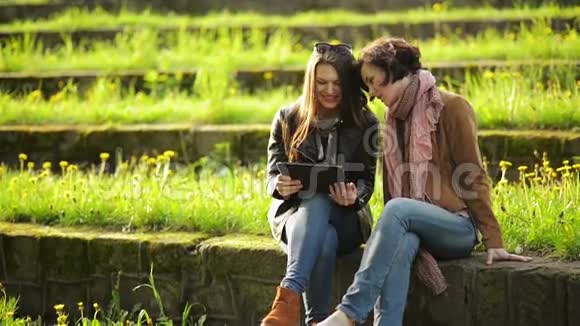 两个漂亮的女孩正在使用坐在公园里的平板户外女朋友正在看电影视频的预览图