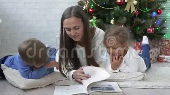 另一位年轻的母亲带着两个孩子在圣诞树附近的地板上看书视频的预览图