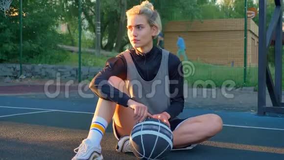 漂亮的金发女篮球运动员坐在地上玩球在后台停车视频的预览图