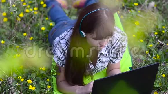 美丽的女孩带着耳机在公园的笔记本电脑上运行女孩在春天公园的电脑上上网聊天视频的预览图