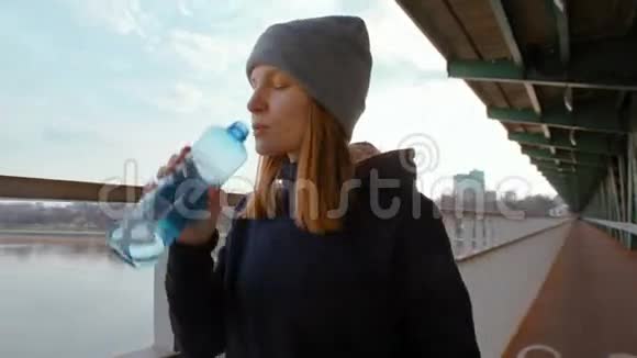 年轻的女士正在河和城市的桥上喝着瓶子里的水视频的预览图