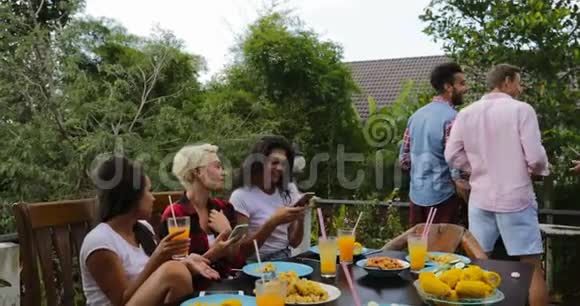 年轻女孩坐在餐桌边聊天而男人集体烹饪烧烤的朋友聚集在夏天的露台上聊天视频的预览图