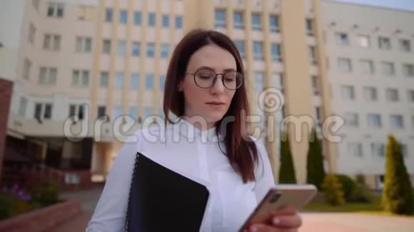 商业女性穿着白色衬衫在城市街道上行走和使用智能手机的正面景色视频的预览图