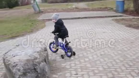 慢动作穿着灰色帽子和温暖夹克的小男孩骑着蓝色自行车在操场上视频的预览图