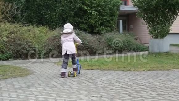 穿着粉红色夹克和帽子的可爱女孩在院子里骑着一辆迷你滑板车踢滑板车视频的预览图