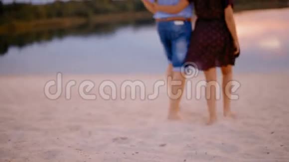 日落时分一对年轻夫妇在沙滩上的沙滩上奔向森林风景如画的湖视频的预览图