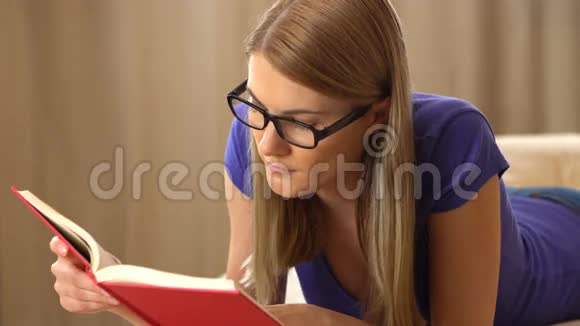 美丽迷人的年轻女人穿着紫罗兰色的t恤和一副黑色眼镜躺在沙发上看书变成一个视频的预览图