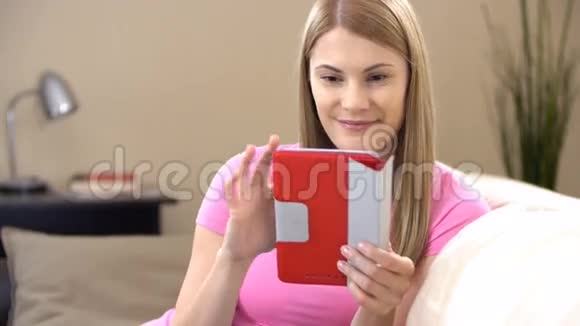 漂亮迷人的年轻女人穿着t恤平板电脑坐在沙发上浏览互联网微笑视频的预览图