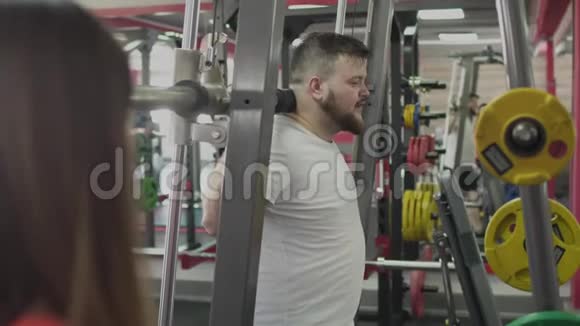 在健身房为大腹便便的男人与女孩教练进行个人培训厚胖的家伙和私人教练一起视频的预览图