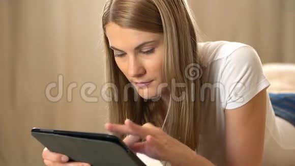 美丽迷人的年轻女子穿着白色t恤一台平板电脑躺在沙发上浏览互联网及视频的预览图