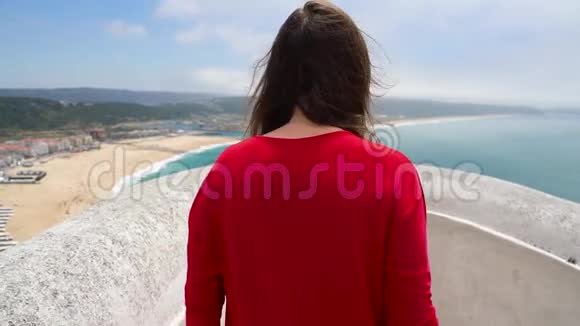 穿着红色衣服的女人可以欣赏到葡萄牙拿撒勒附近的海洋海岸慢动作视频的预览图