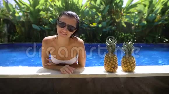 在炎热的天气里美丽的女孩正在游泳池里靠在舞台上那里有两个菠萝在一个舞台上视频的预览图